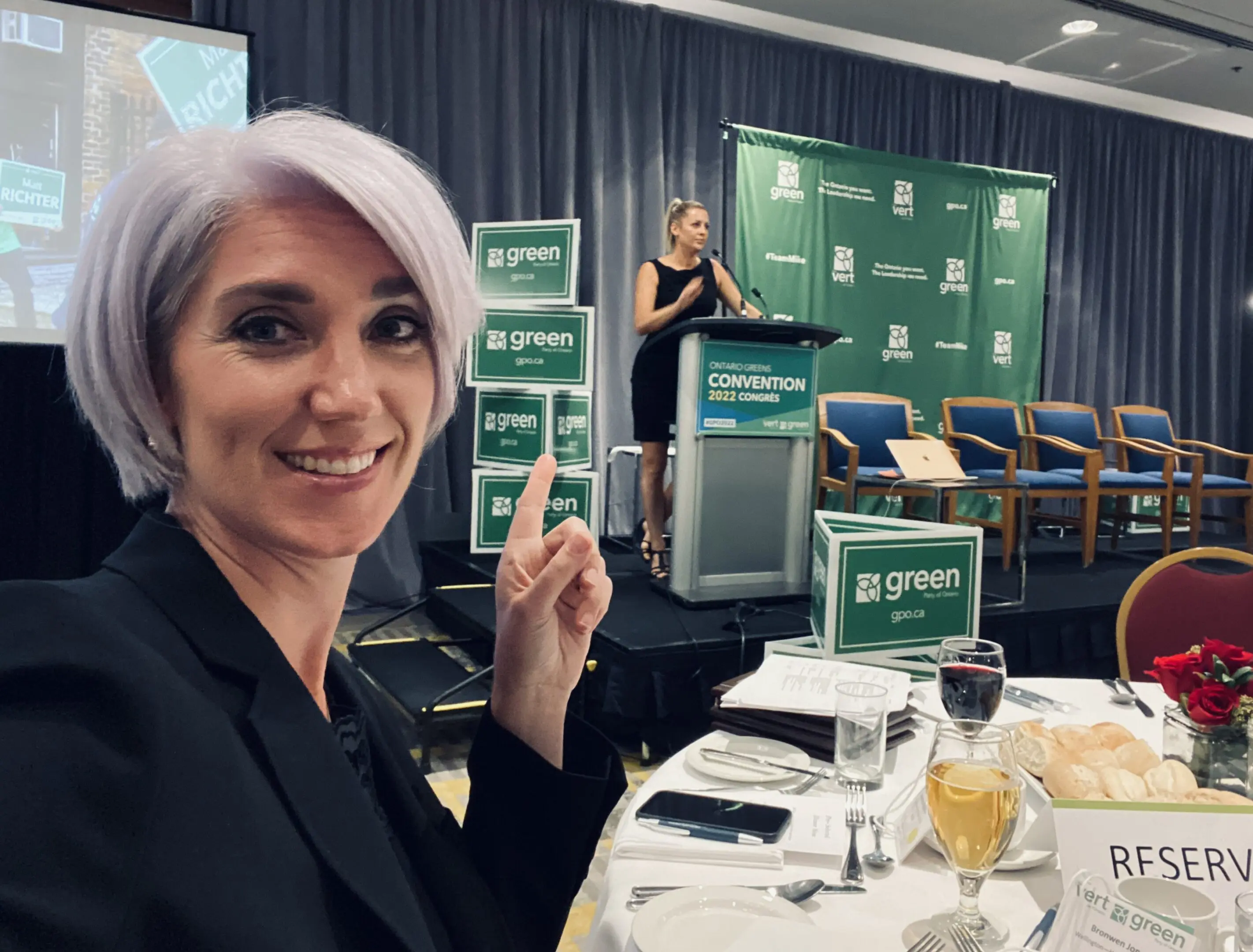 Woman with silver hair taking a selfie at a formal event with a speaker in the background.